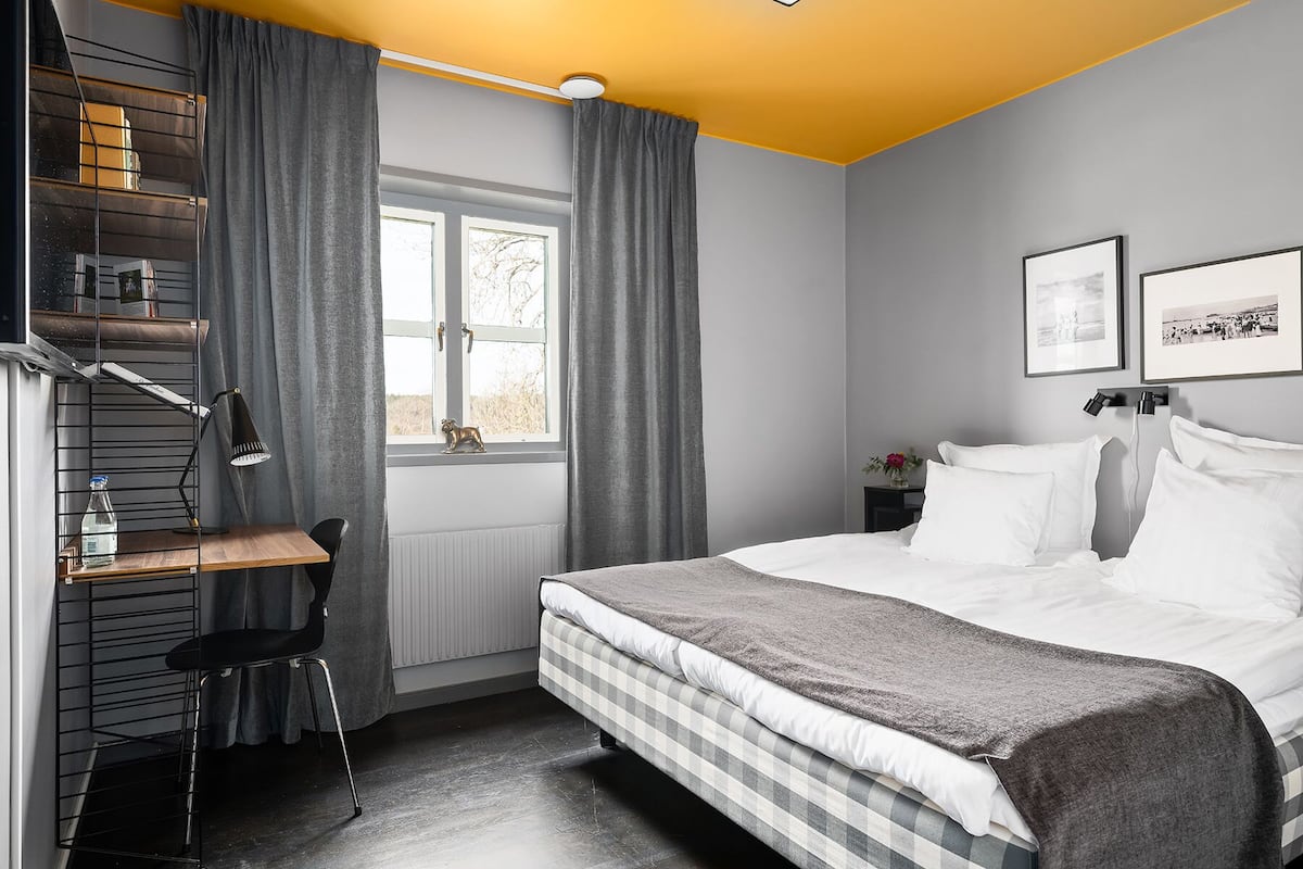 A bedroom with a bed, a desk with a chair, a radiator, a window with curtains, and a wall with framed pictures.