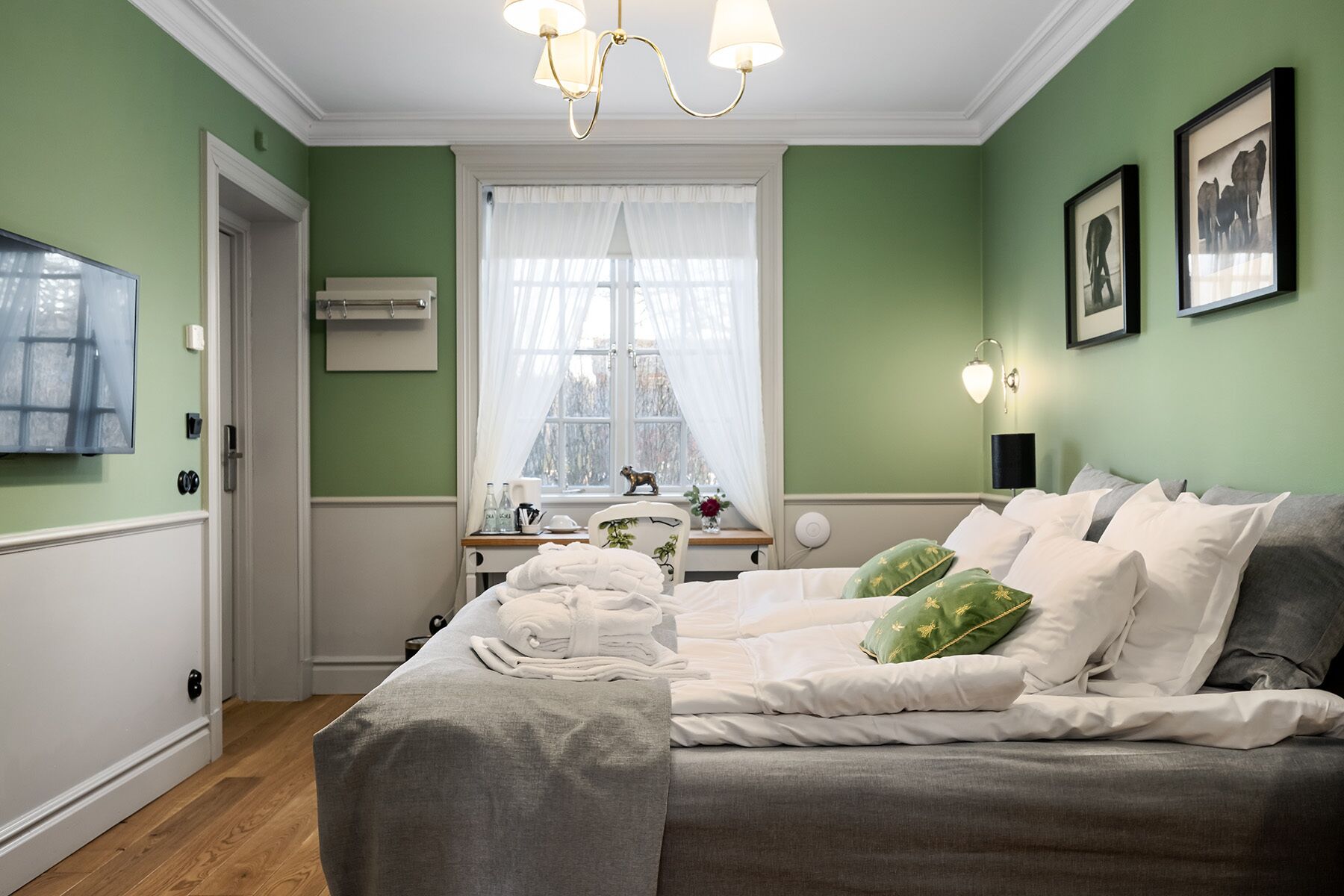 Un dormitorio con cama, ventana con cortinas, mesita de noche con lámpara y televisor montado en la pared.
