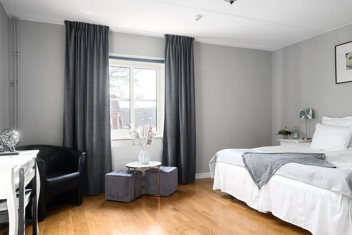 A modern bedroom with a large window, a bed, a chair, and a small table with a vase.