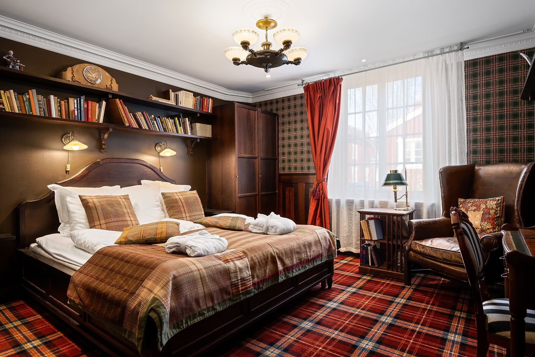 Un dormitorio tradicional con una cama grande, cabecera de madera, estantería, alfombra de cuadros rojos y un candelabro clásico.