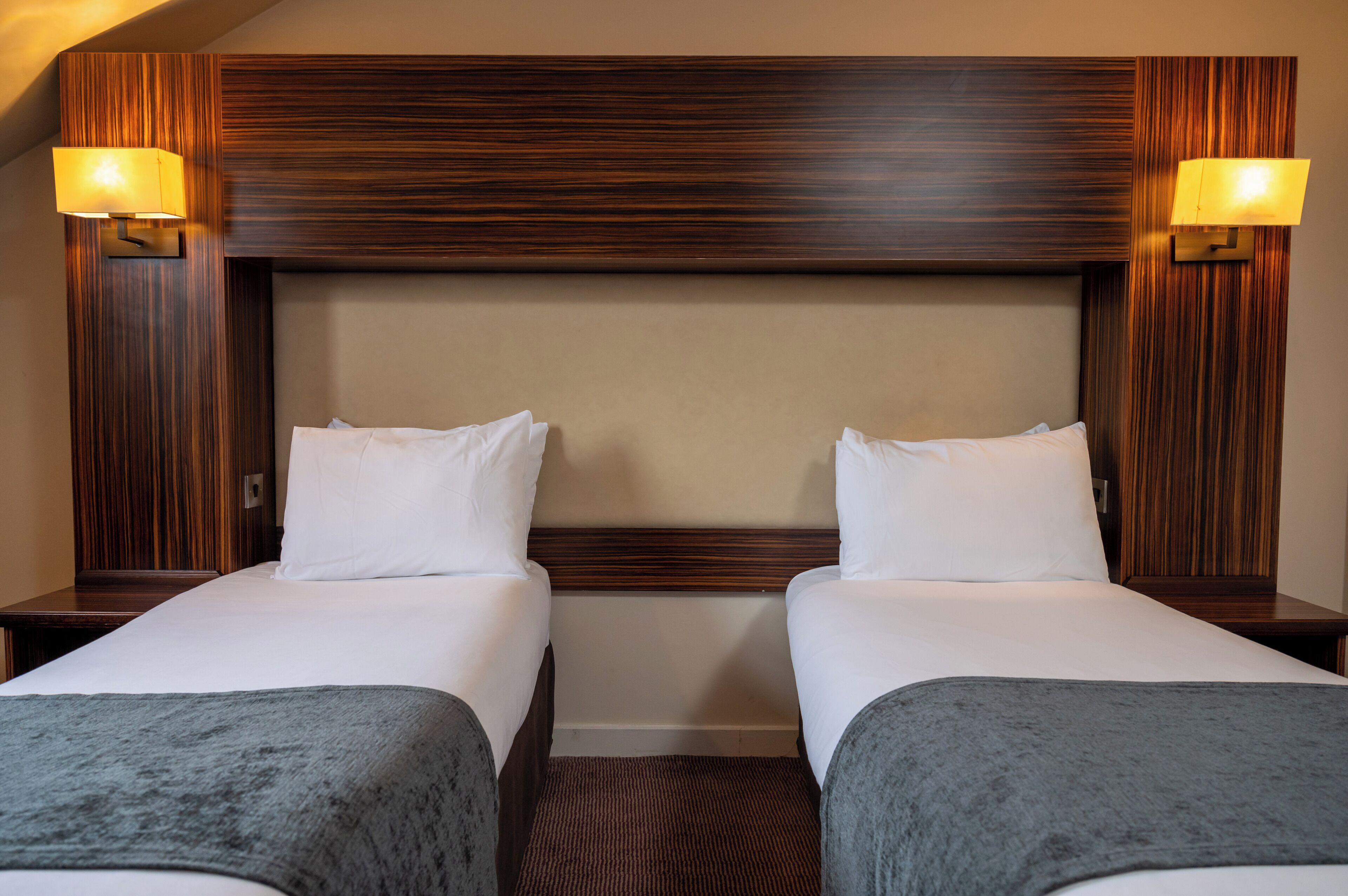 A hotel room with two beds, wooden headboard, and two wall-mounted lamps.