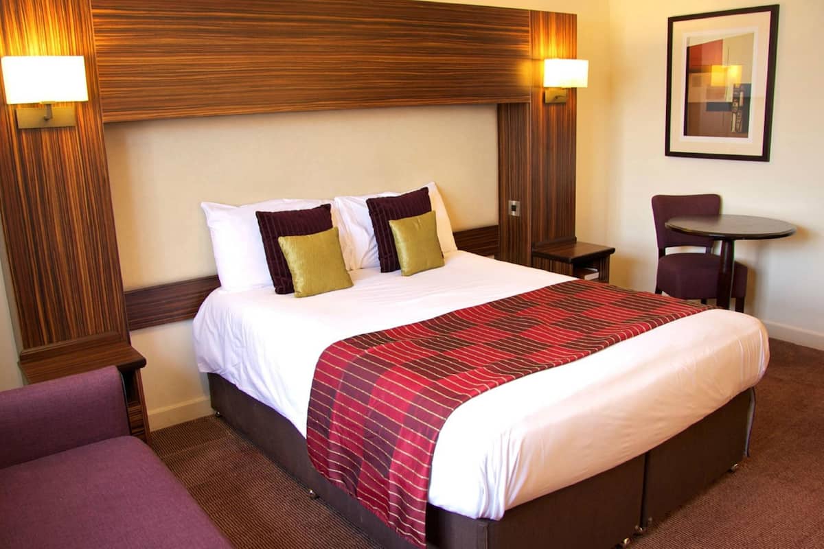 A hotel room with a bed, a chair, a small table, and a wall-mounted lamp.