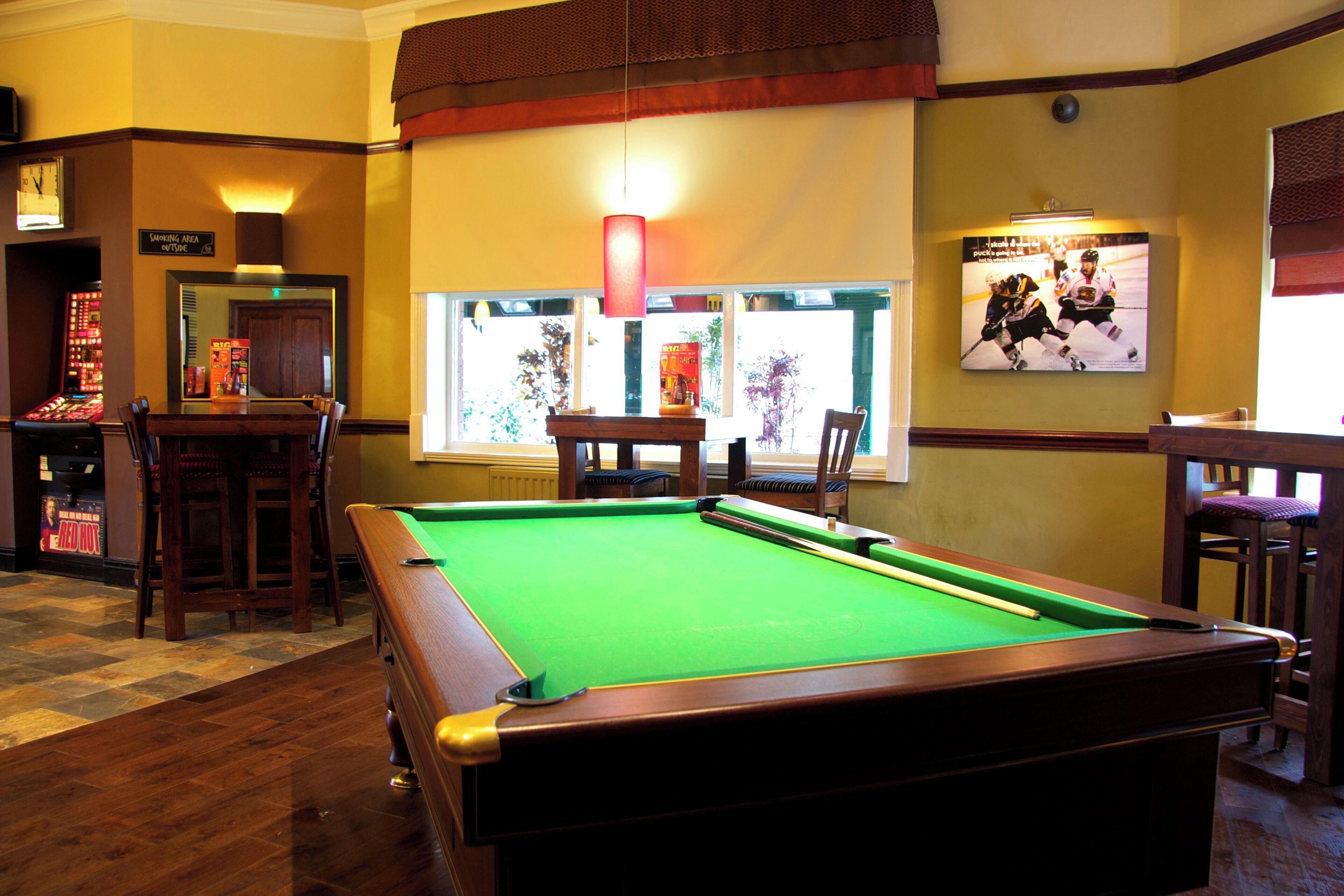 A pool table in a room with wooden flooring, a bar area with stools, and a television displaying a hockey game.