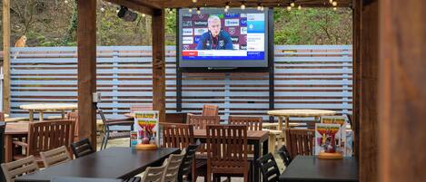 An outdoor seating area with wooden tables and chairs, a TV screen displaying a sports broadcast, and a wooden fence in the background.