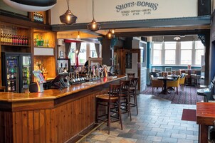 Zona de bar con mostrador de madera, taburetes y una variedad de botellas y vasos. Hay un cartel con la carta sobre el mostrador y un área de estar con mesas y sillas al fondo.