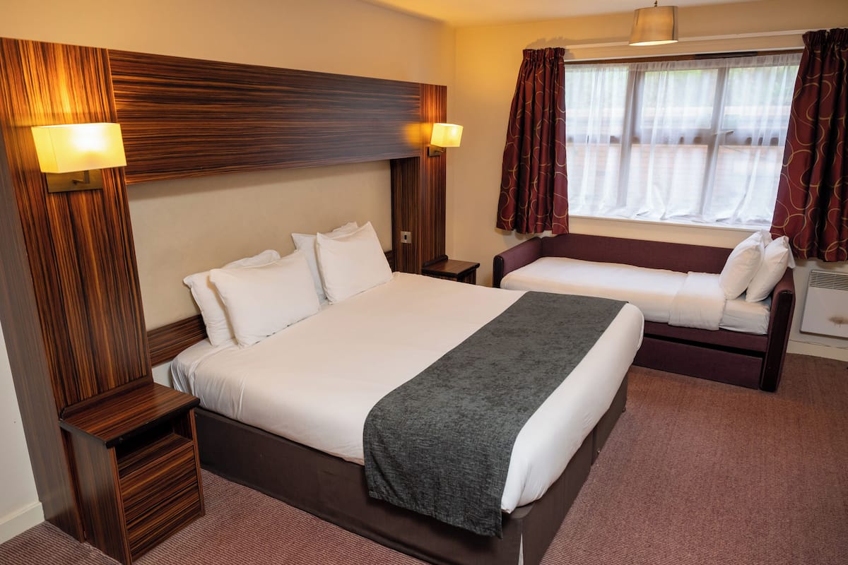 A hotel room with a large bed, a smaller bed, a nightstand, a lamp, a window with curtains, and a wooden headboard.