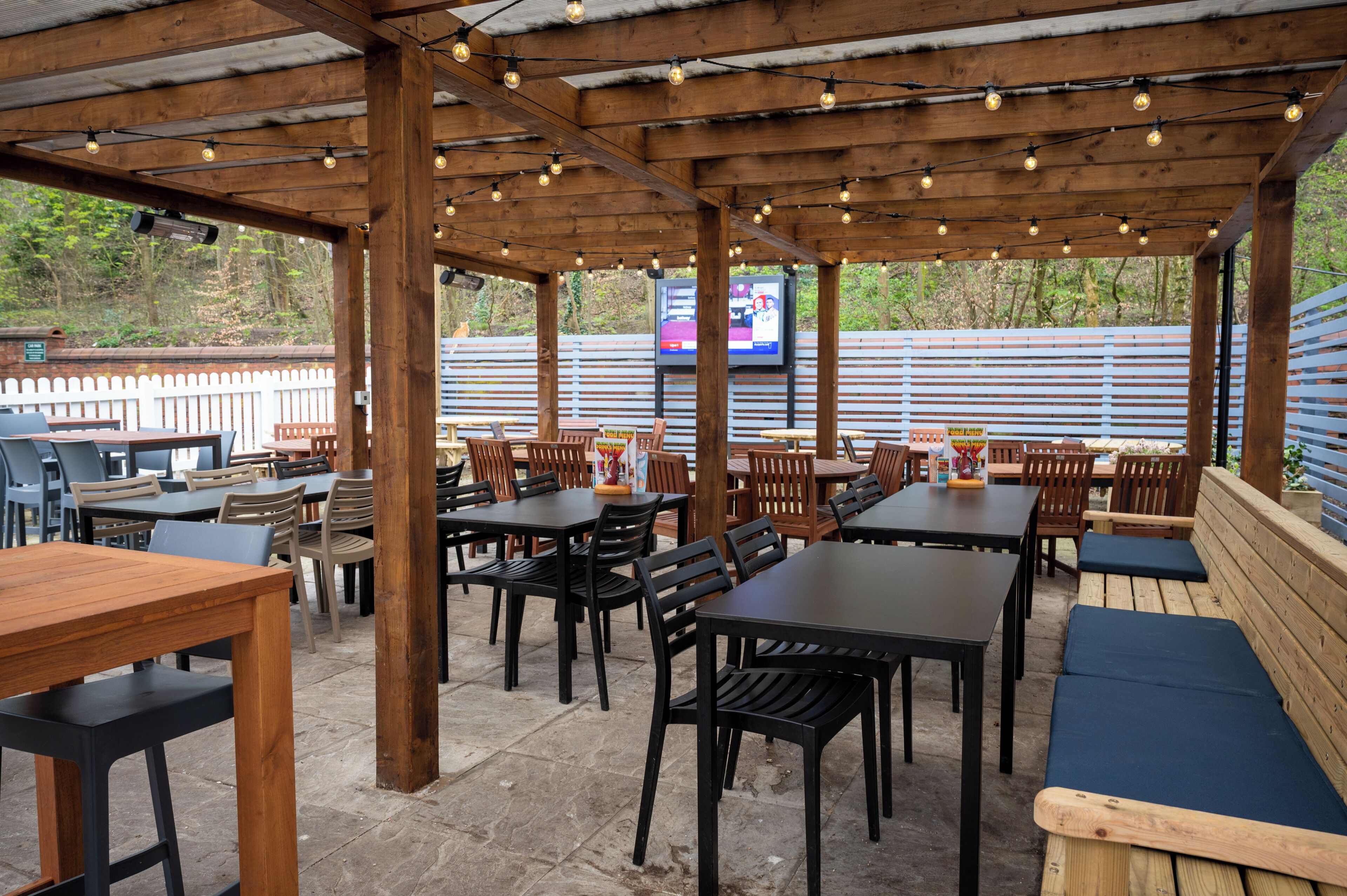 An outdoor patio area with wooden tables and chairs, a TV screen, and a white picket fence in the background.