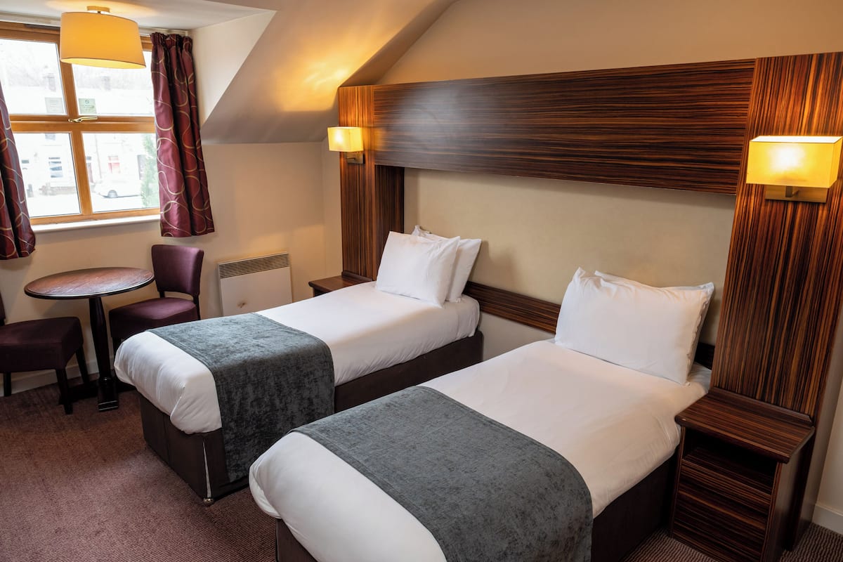 A hotel room with two single beds, a small round table, a chair, and a window with curtains.