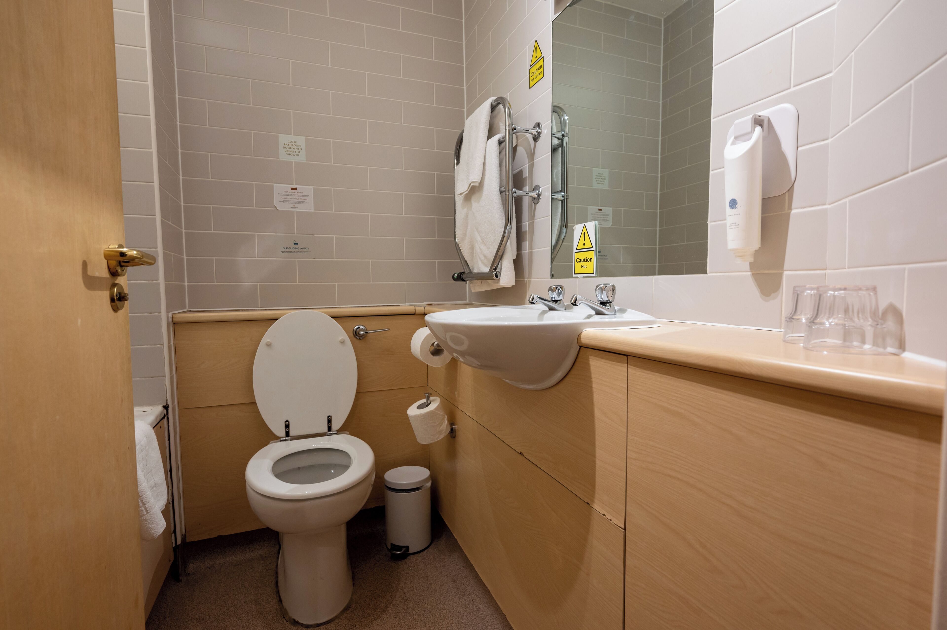 A small bathroom with a toilet, sink, and mirror. There are towels hanging on the wall and a trash can.