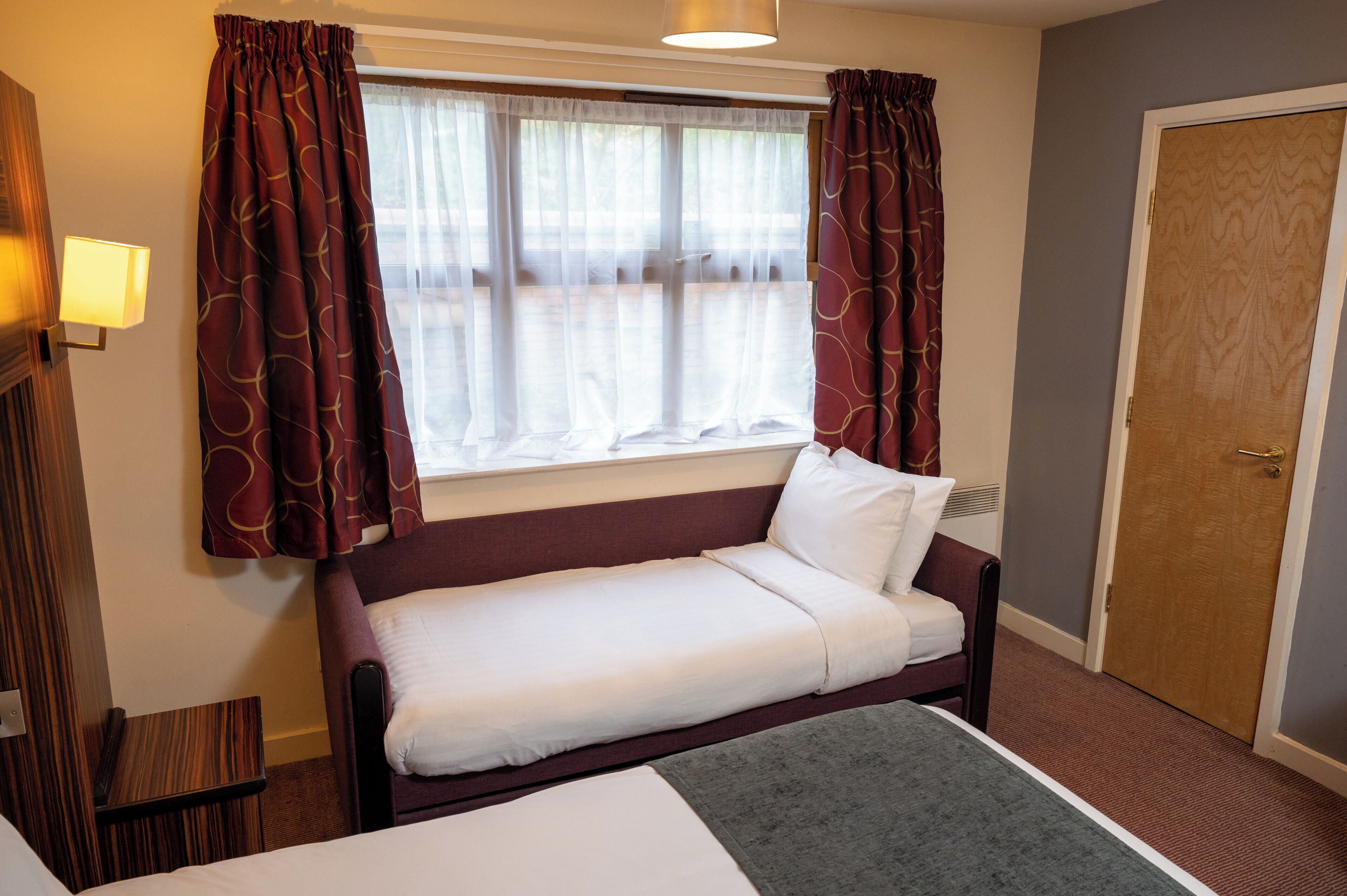 A single bed with a grey blanket, a small bedside table, a window with sheer curtains, and a door to the right.