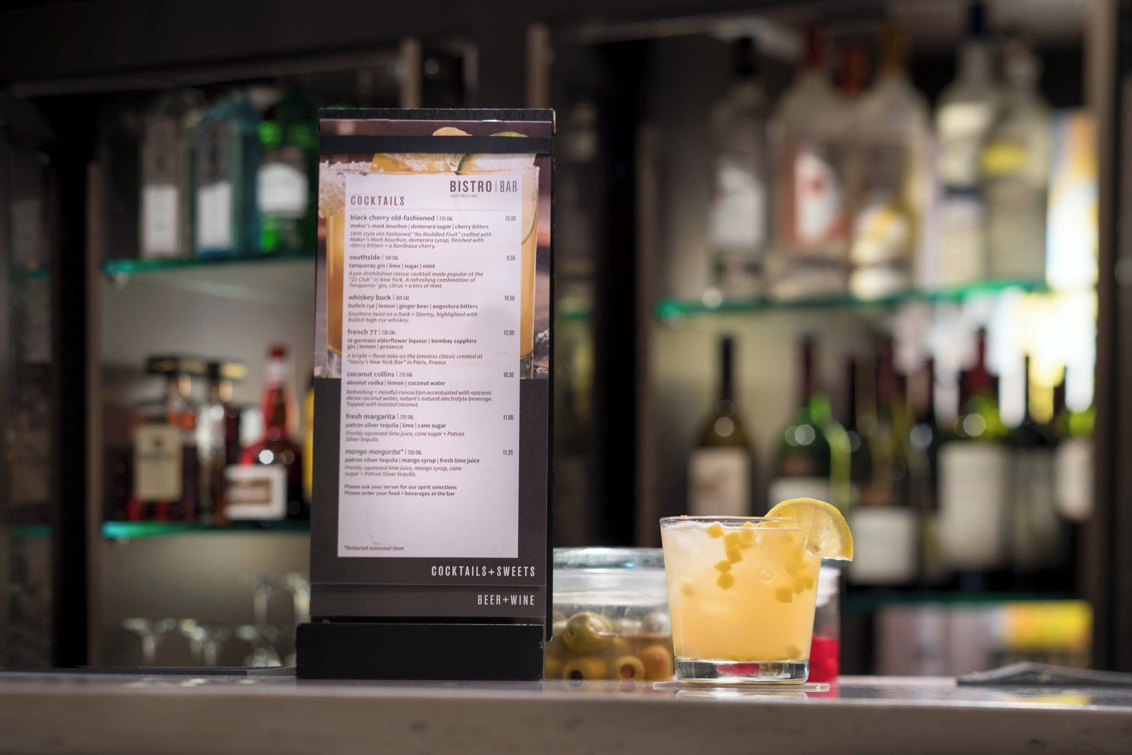 A bar menu with cocktails and sweets, and a glass of cocktail with a lemon slice on a bar counter.