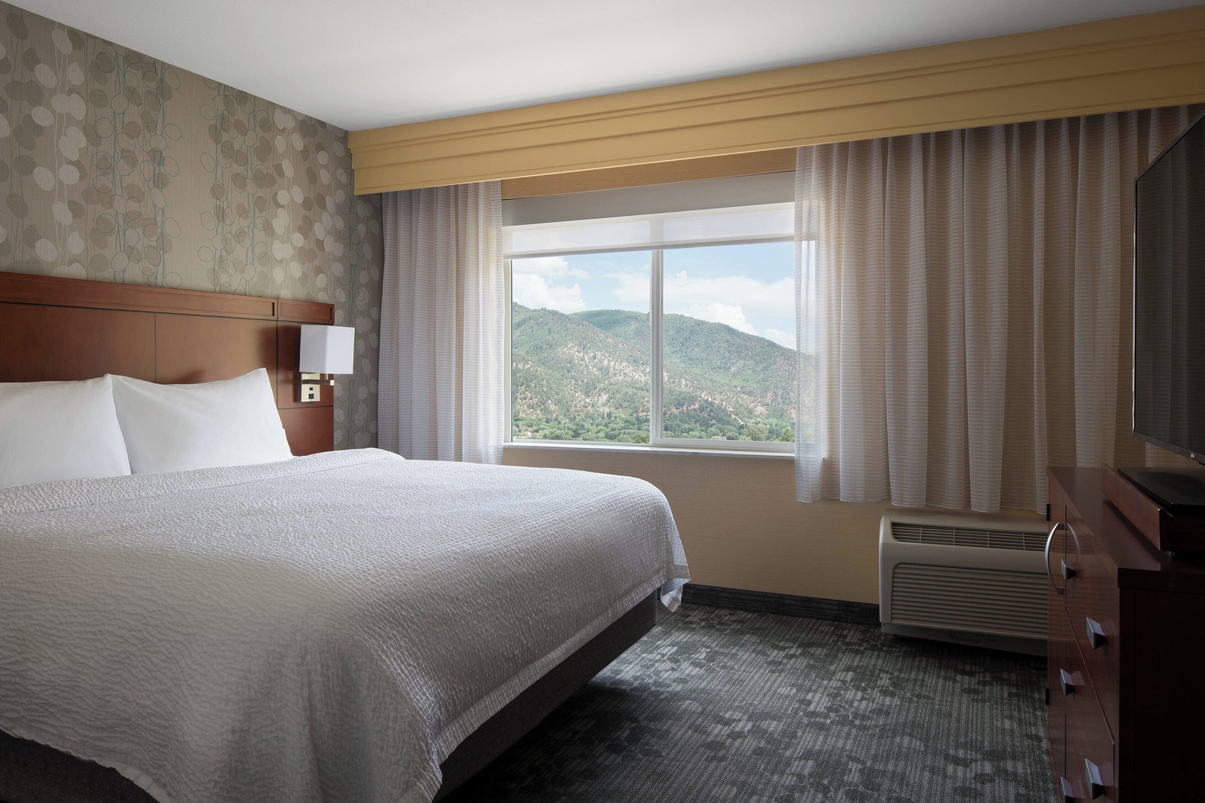 Habitación de hotel con una cama grande, una mesita de noche con lámpara, un aparador y una ventana con vistas a un paisaje montañoso.