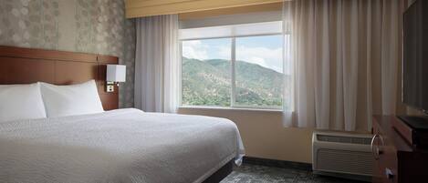 Habitación de hotel con una cama grande, una mesita de noche con lámpara, un aparador y una ventana con vistas a un paisaje montañoso.