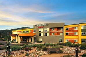 Un hôtel Courtyard Marriott au design moderne, caractérisé par un mélange de bâtiments aux couleurs jaune et grise, un jardin aménagé avec un sentier et une statue en premier plan.