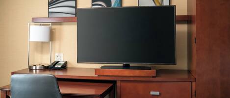 A hotel room with a large flat-screen TV, a desk with a telephone, and a chair. There are three framed artworks on the wall above the TV.
