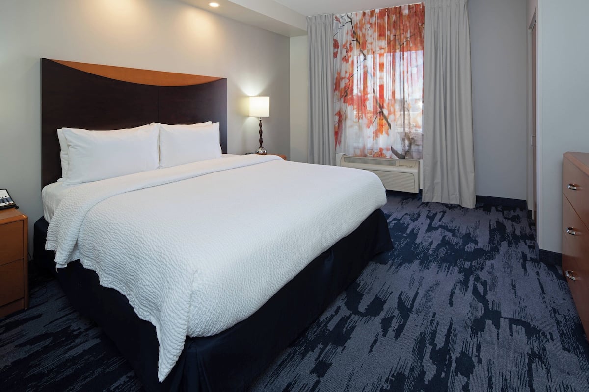 Une chambre d’hôtel avec un grand lit, une table de chevet, une lampe, une fenêtre avec des rideaux et une commode.