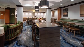 A hotel lobby with a central bar, seating areas, and a kitchen in the background.