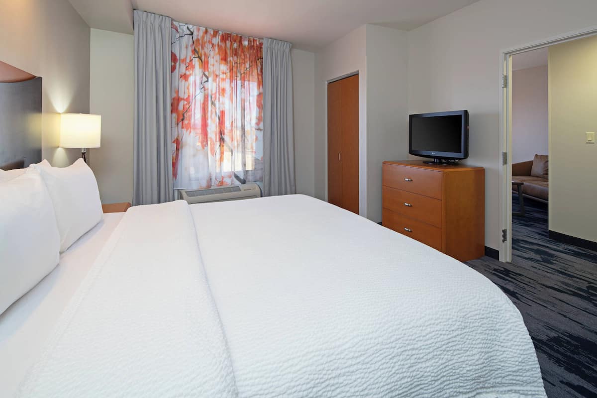Une chambre d’hôtel avec un grand lit, un téléviseur posé sur une commode en bois, et une vue sur une autre chambre par une porte ouverte.