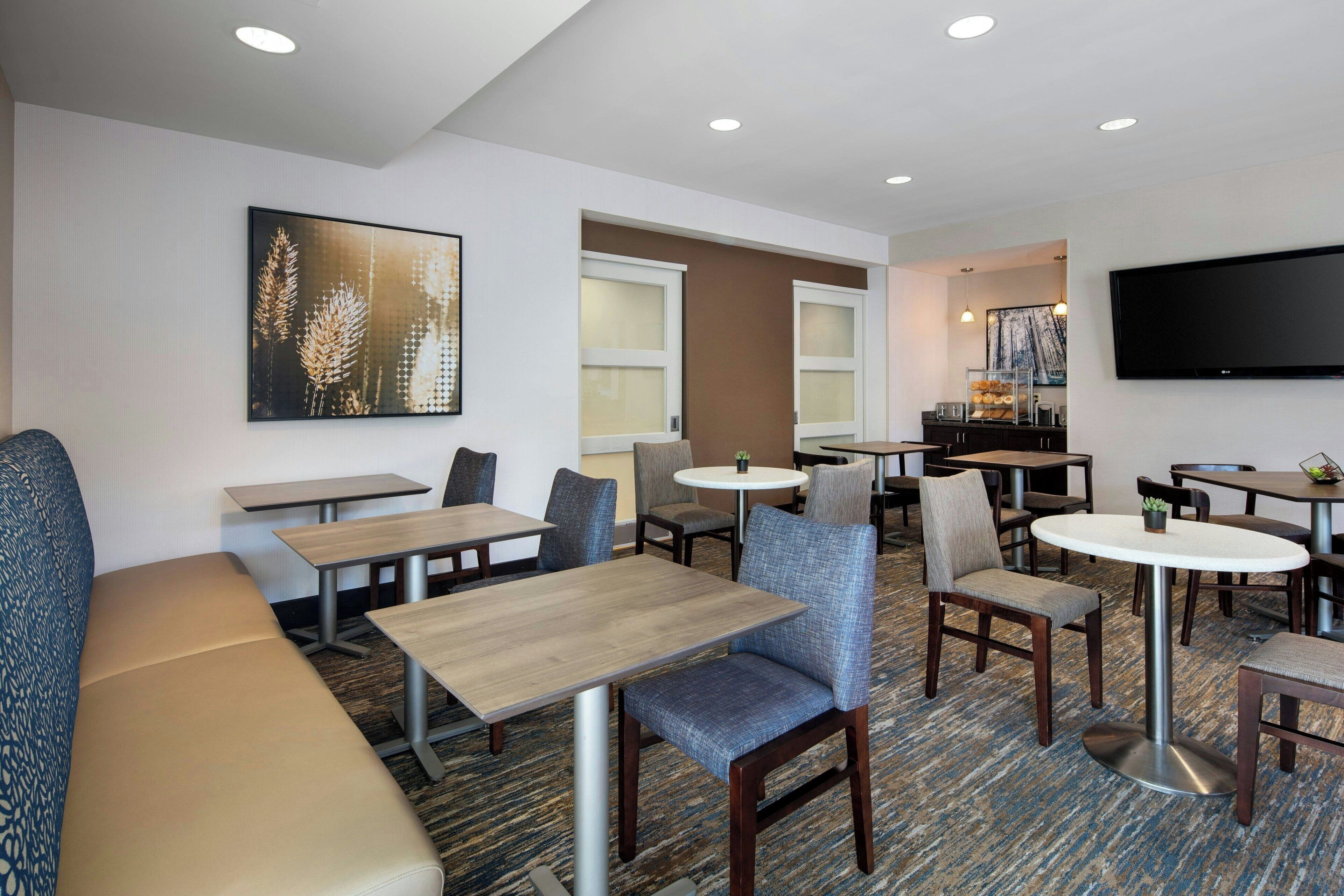 A hotel lobby with multiple seating areas, including sofas and tables, a flat-screen TV mounted on the wall, and a large window providing natural light.