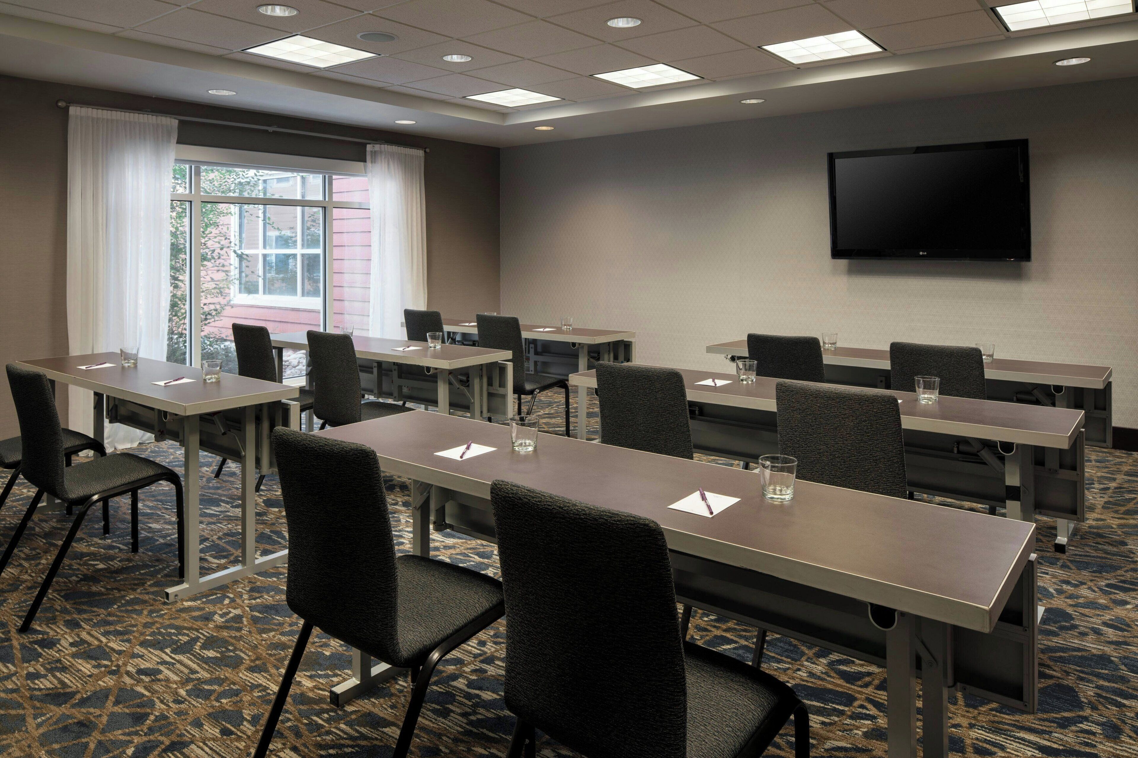 A conference room with multiple tables and chairs, a flat-screen TV mounted on the wall, and a large window with curtains.