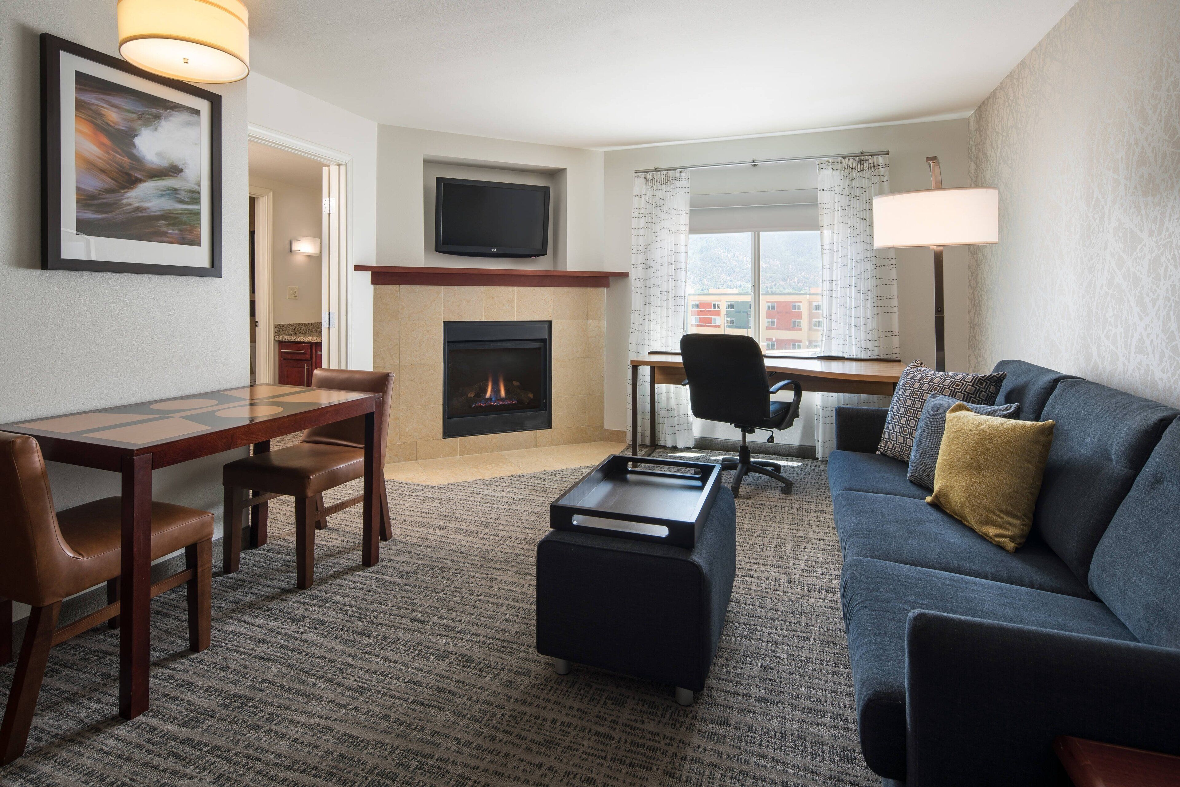 A modern living room with a fireplace, a flat-screen TV, a dining area with a table and chairs, and a sofa with a patterned throw pillow.