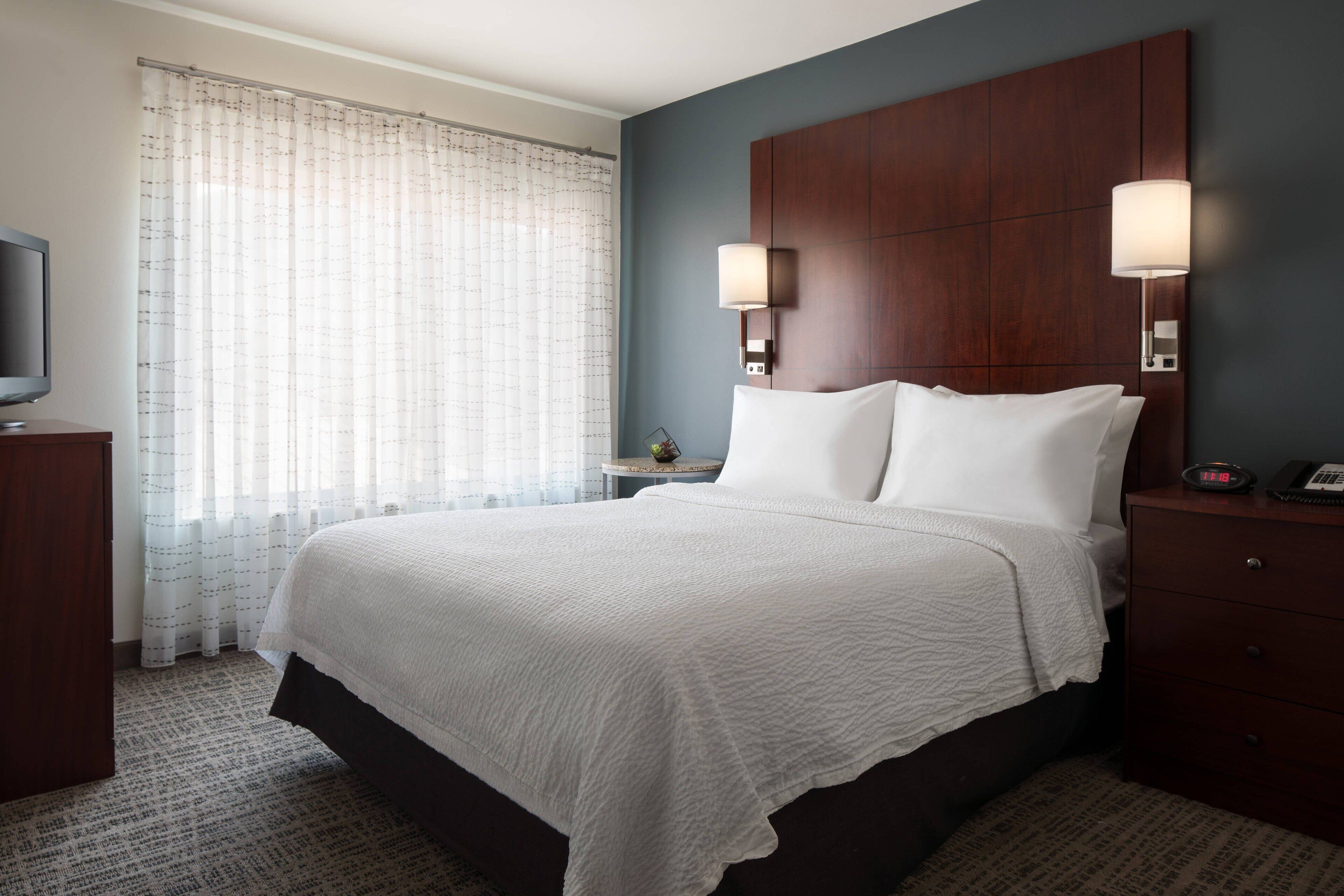 A hotel room with a large bed, a wooden headboard, two bedside lamps, a nightstand, a television, and a window with sheer curtains.