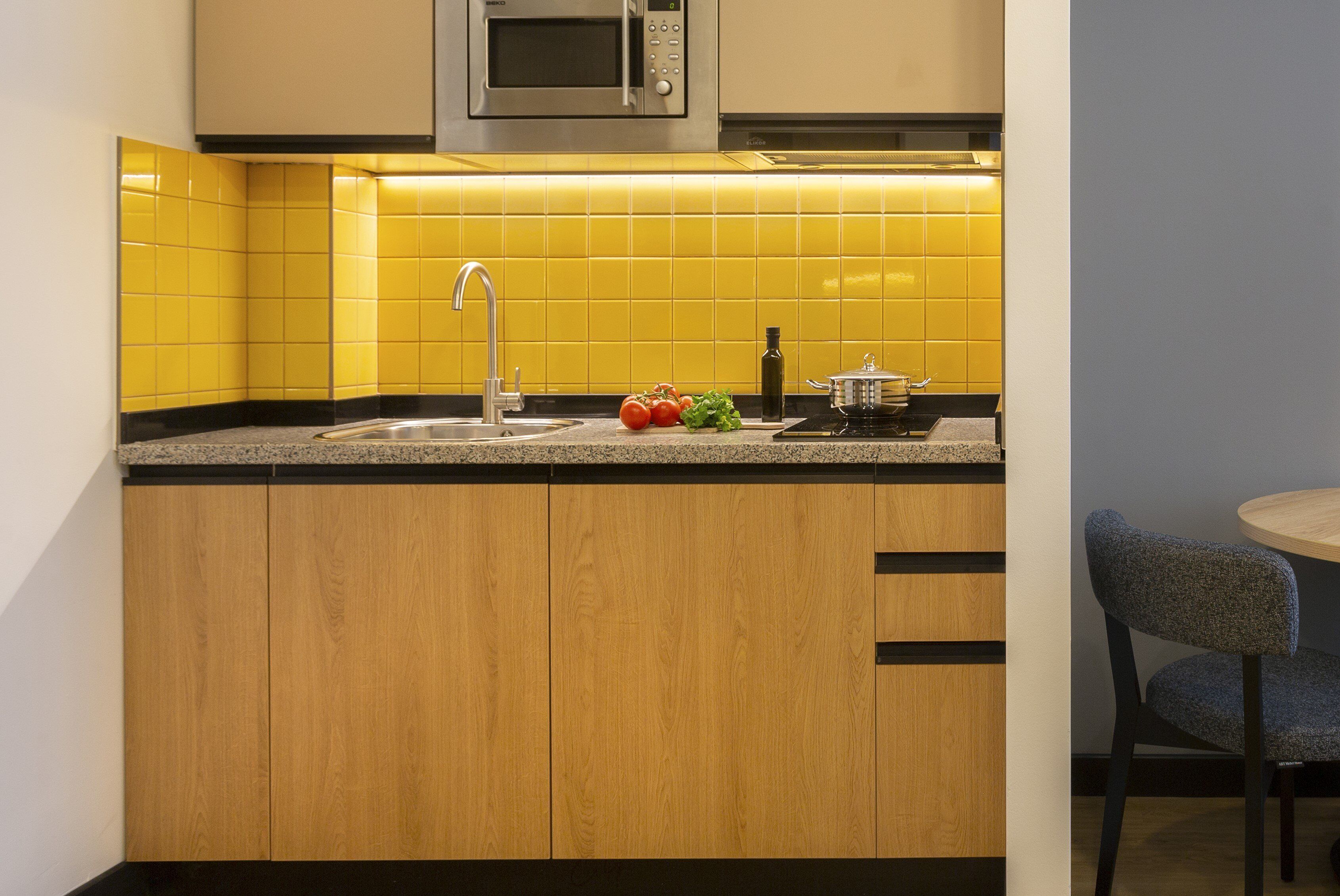 A compact kitchen with yellow tiles, a stainless steel microwave, a sink, and a small dining area with a chair.