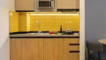 A compact kitchen with yellow tiles, a stainless steel microwave, a sink, and a small dining area with a chair.