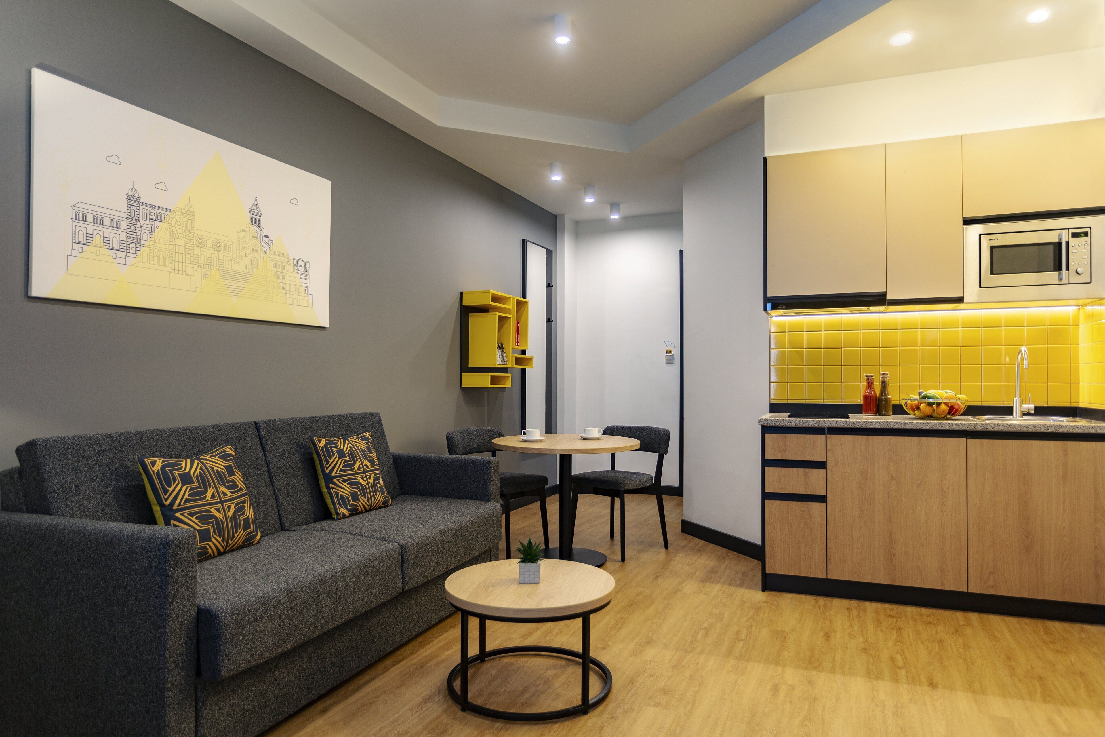 A modern living room with a grey sofa, a small round table, and a kitchen area with yellow tiles and wooden cabinets.
