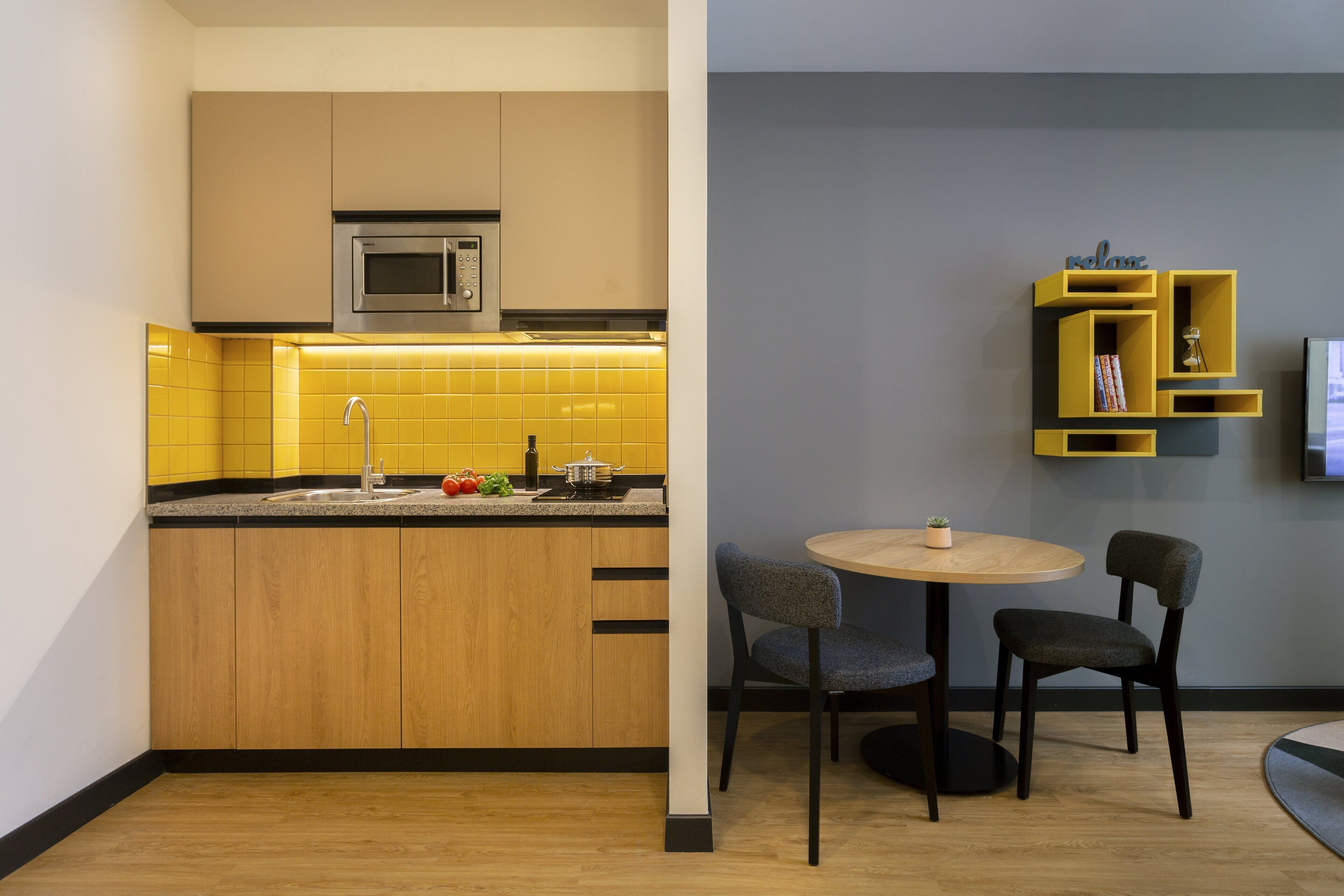 Una cocina moderna con armarios de madera, microondas integrado y salpicadero con azulejos amarillos. Hay un comedor con una mesa redonda y dos sillas.