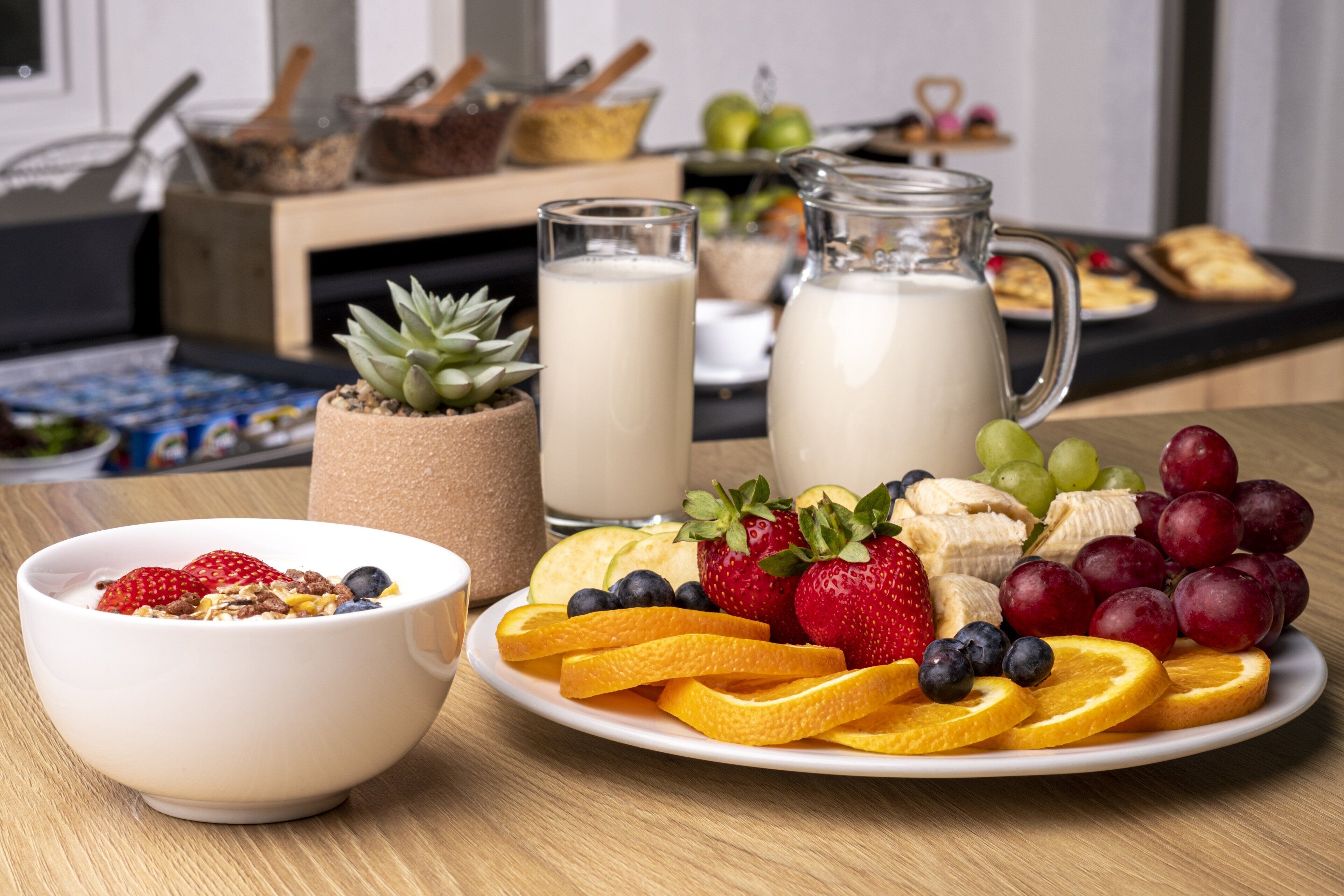 Un desayuno con un tazón de cereal, un plato de fruta, un vaso de leche y una jarra de leche.