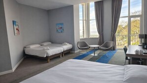 Habitación de hotel con una cama grande, dos camas más pequeñas, un escritorio con una silla, una mesita con dos sillas y un balcón con barandilla.