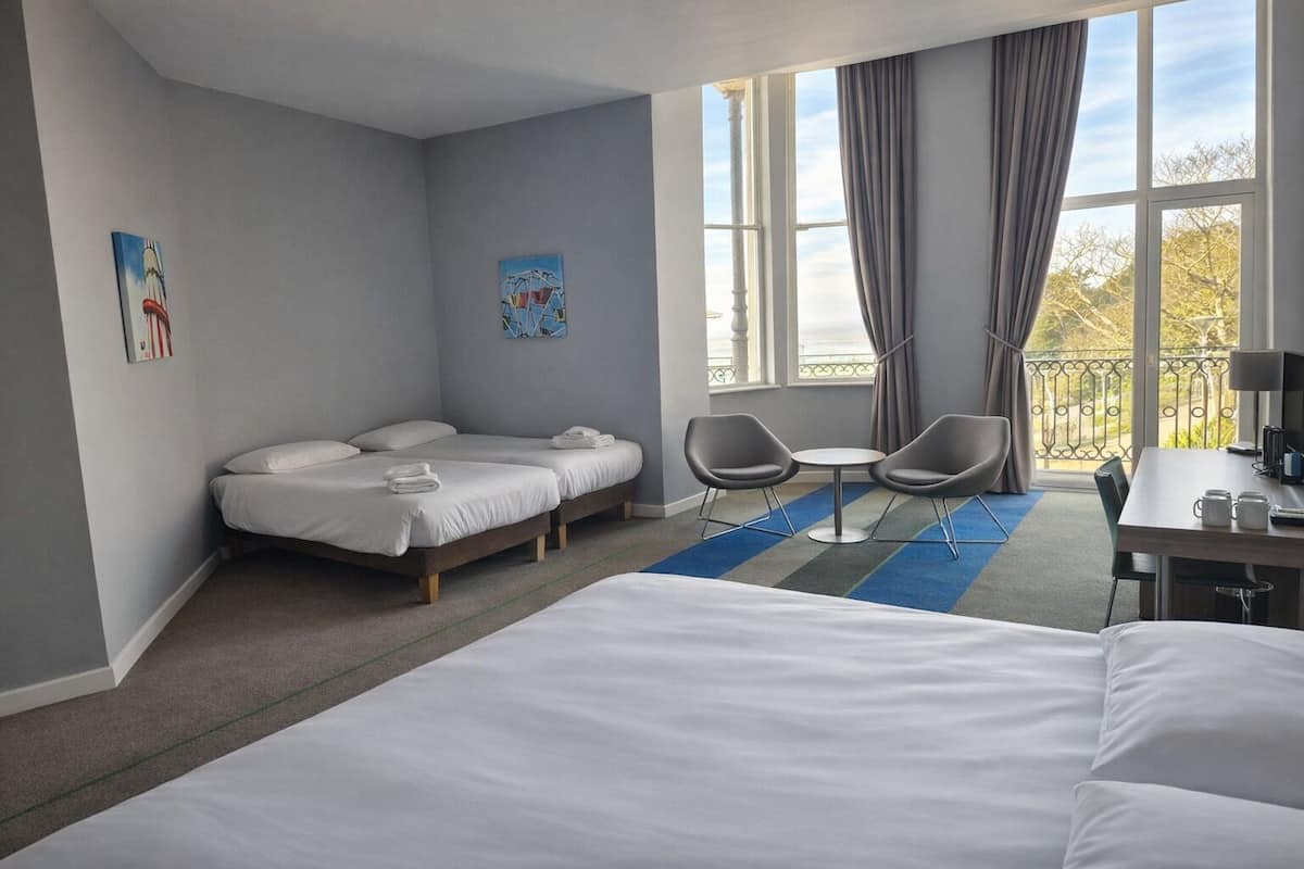 Habitación de hotel con una cama grande, dos camas más pequeñas, un escritorio con una silla, una mesita con dos sillas y un balcón con barandilla.