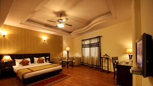 A hotel room with a large bed, two bedside tables with lamps, a ceiling fan, a TV, and a small table with a chair. - Hotel The Class (New Delhi)