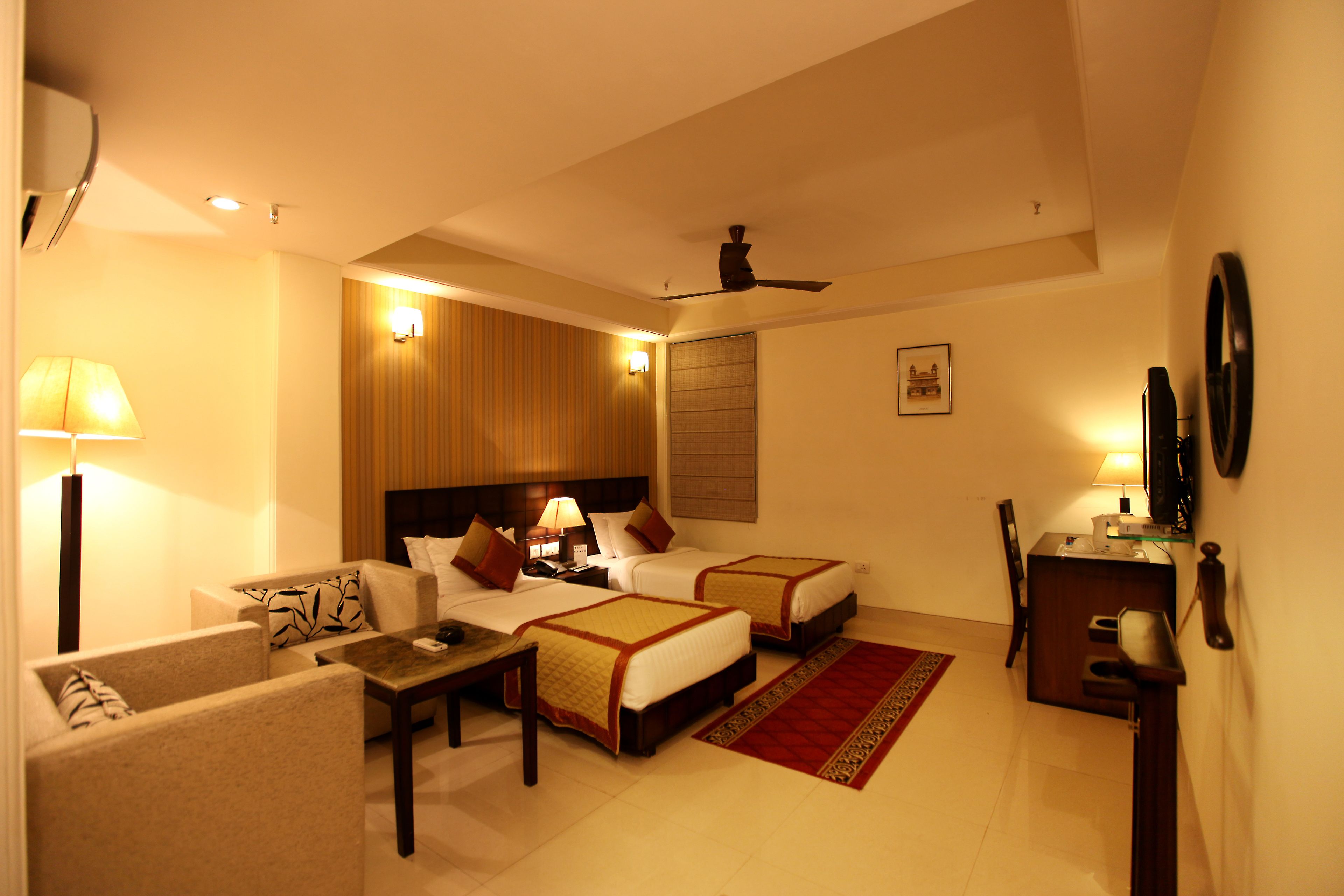 A hotel room with two beds, a desk, a chair, a lamp, and a ceiling fan.