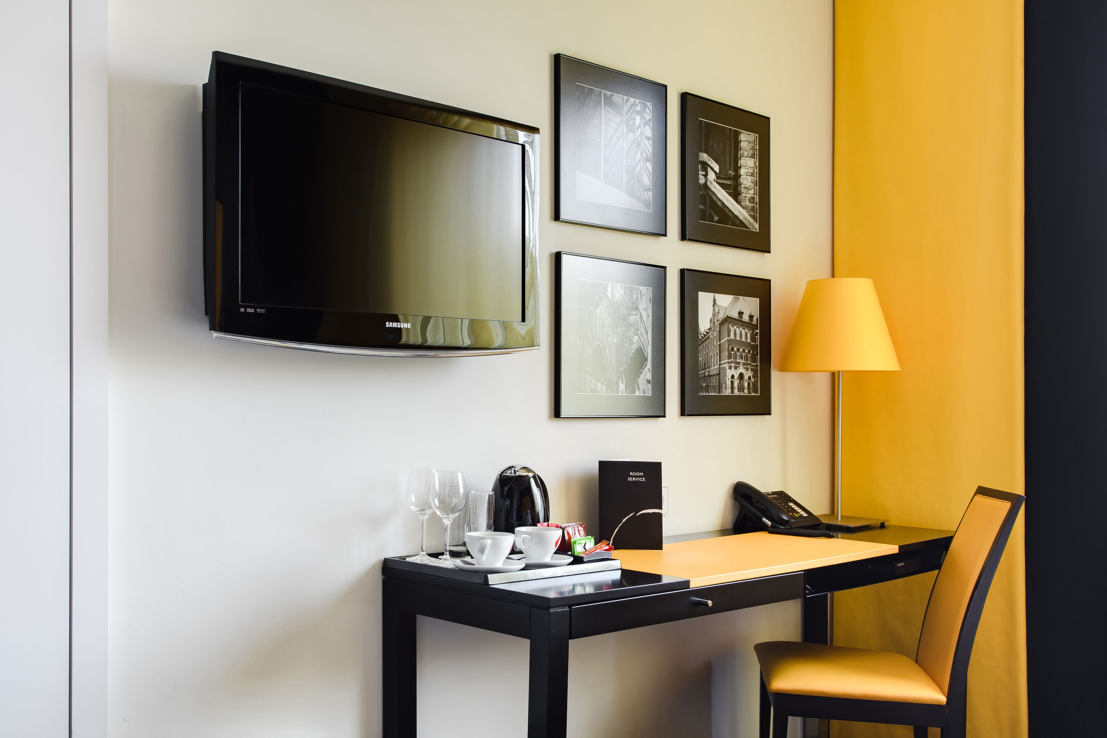 Une chambre d’hôtel équipée d’un téléviseur à écran plat, de tableaux encadrés au mur, d’un bureau avec une lampe, d’une chaise et d’une machine à café.