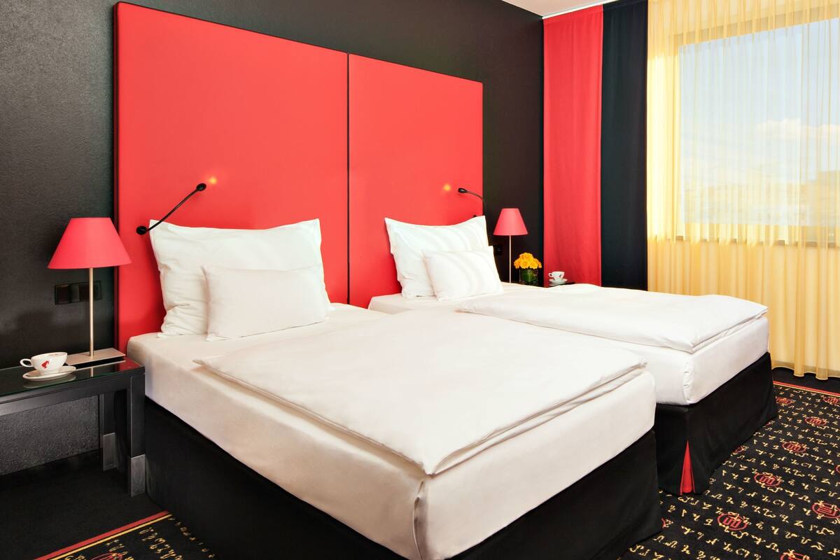 Habitación de hotel con dos camas, una pared de acento roja, una mesita de noche con una lámpara y una ventana con cortinas.