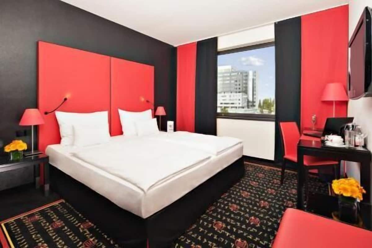 Habitación de hotel con una cama grande, un escritorio con silla, una ventana con vistas a edificios y una decoración en tonos rojo y negro.
