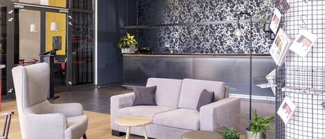 Un espace d’accueil moderne avec un canapé gris, une chaise en bois, une petite table ronde et un présentoir à plantes en métal.