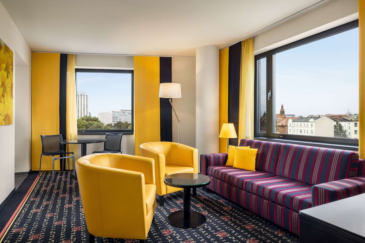 A hotel room with yellow chairs, a striped sofa, a small table, and large windows offering city views.