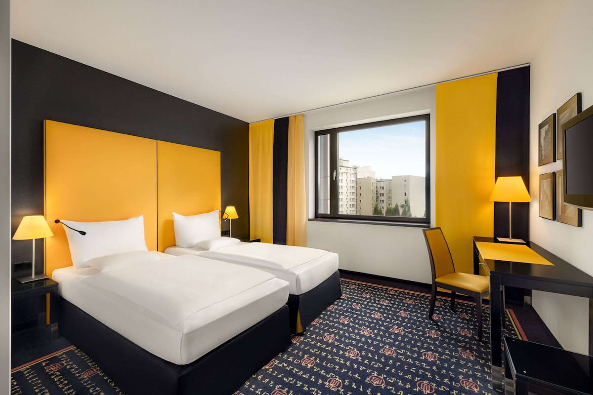 A hotel room with two beds, a desk, a chair, a window with a city view, and a patterned carpet.