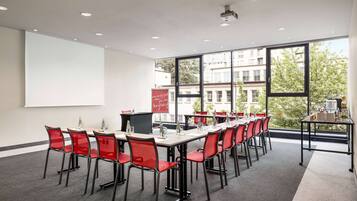A conference room with a long table, red chairs, a projector screen, and large windows with a view of buildings and trees.