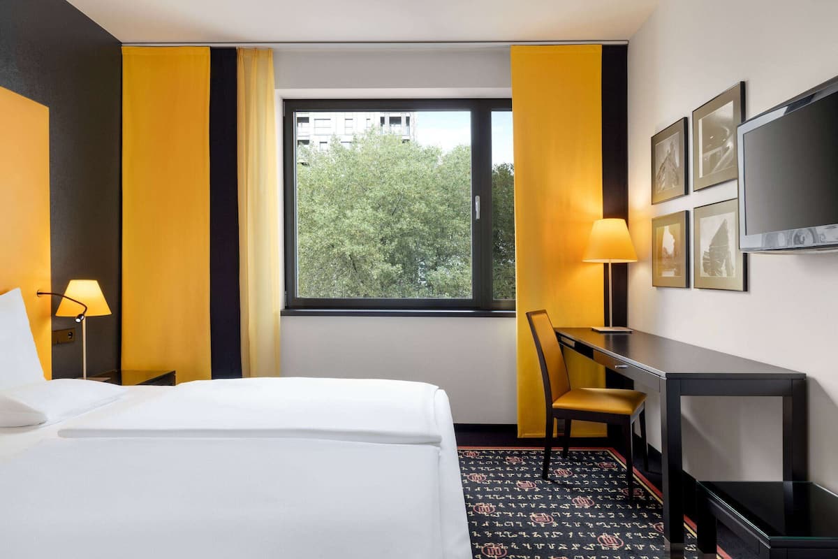 A hotel room with a bed, a desk, a chair, a TV, and a window with a view of trees.