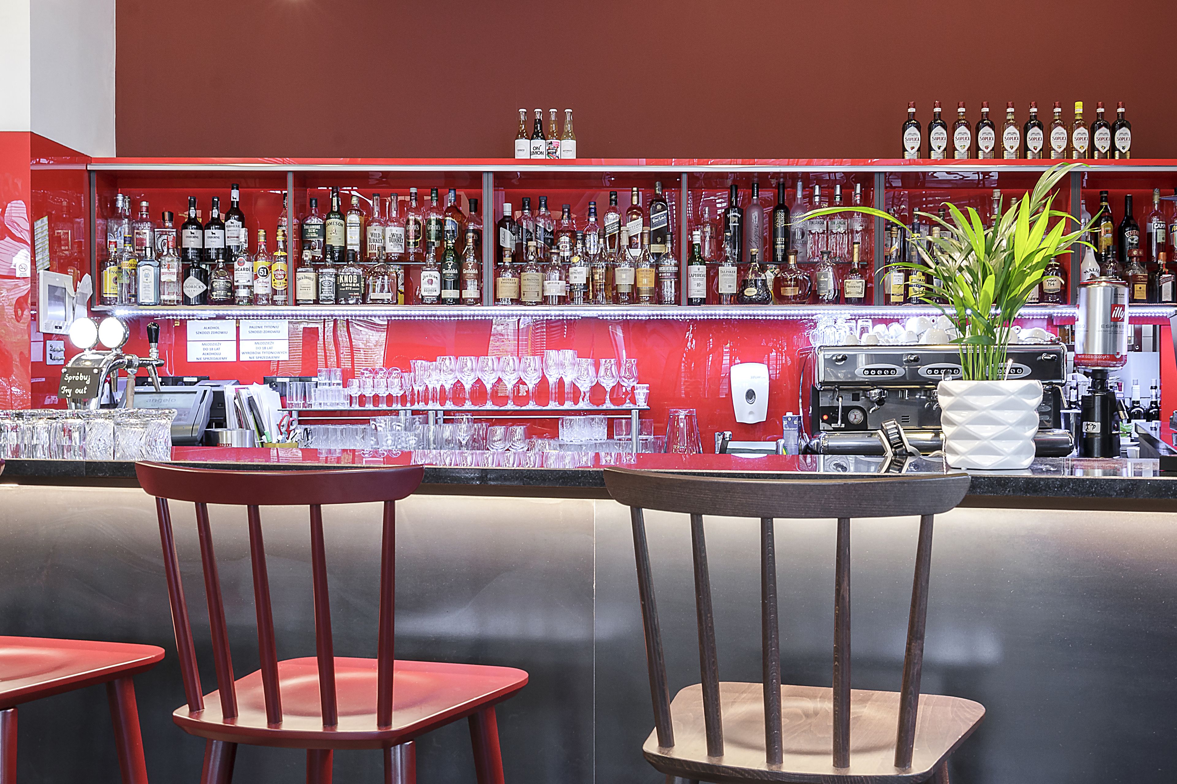 Zona de bar con varias botellas de licor, vasos y una máquina de café. Hay dos sillas de madera y una planta en maceta.