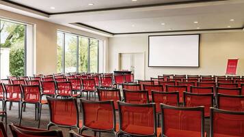 Un salón de conferencias con filas de sillas rojas, una pantalla de proyector y amplias ventanas.