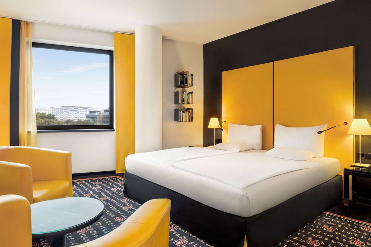 A hotel room with a large bed, a round table, a bookshelf, and a view of the city through the window.