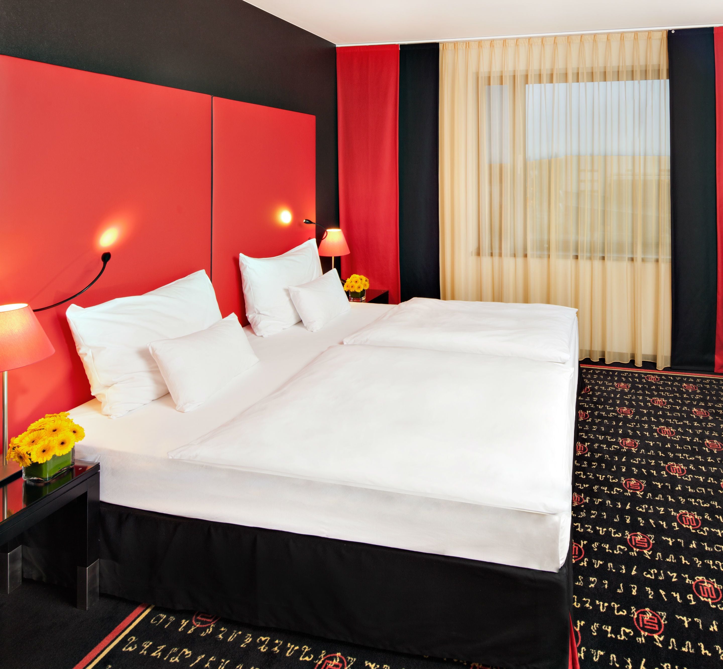 Une chambre d’hôtel avec un grand lit, deux tables de chevet, une petite table, une lampe et une fenêtre avec des rideaux légers.