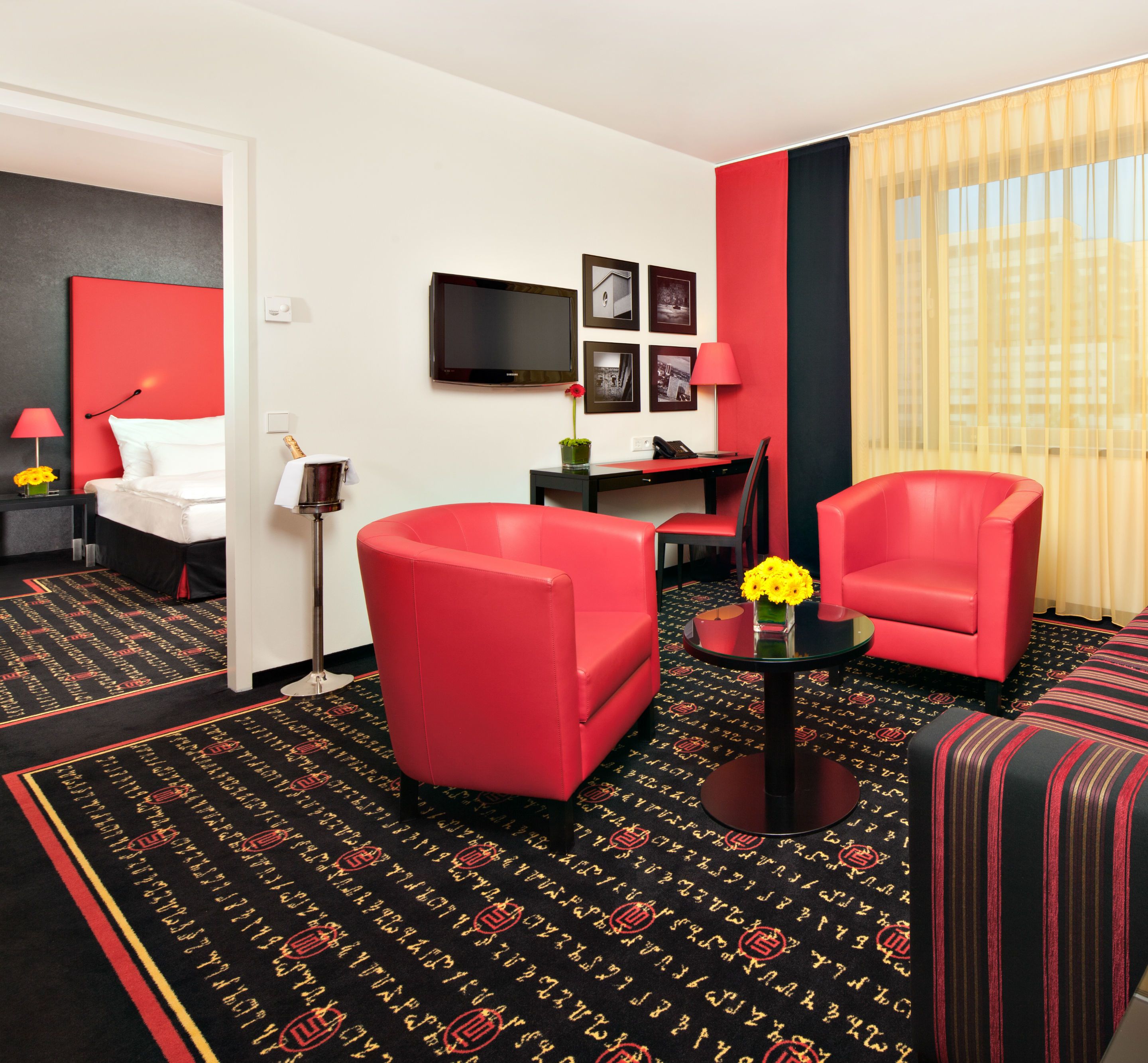 A hotel room with a red and black carpet, a bed, a desk, a chair, a TV, and a window with curtains.