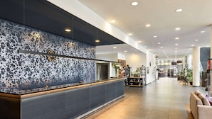 Un espace d’accueil moderne avec un mur à texture, un comptoir de réception et un hall spacieux agrémenté de sièges et de plantes.