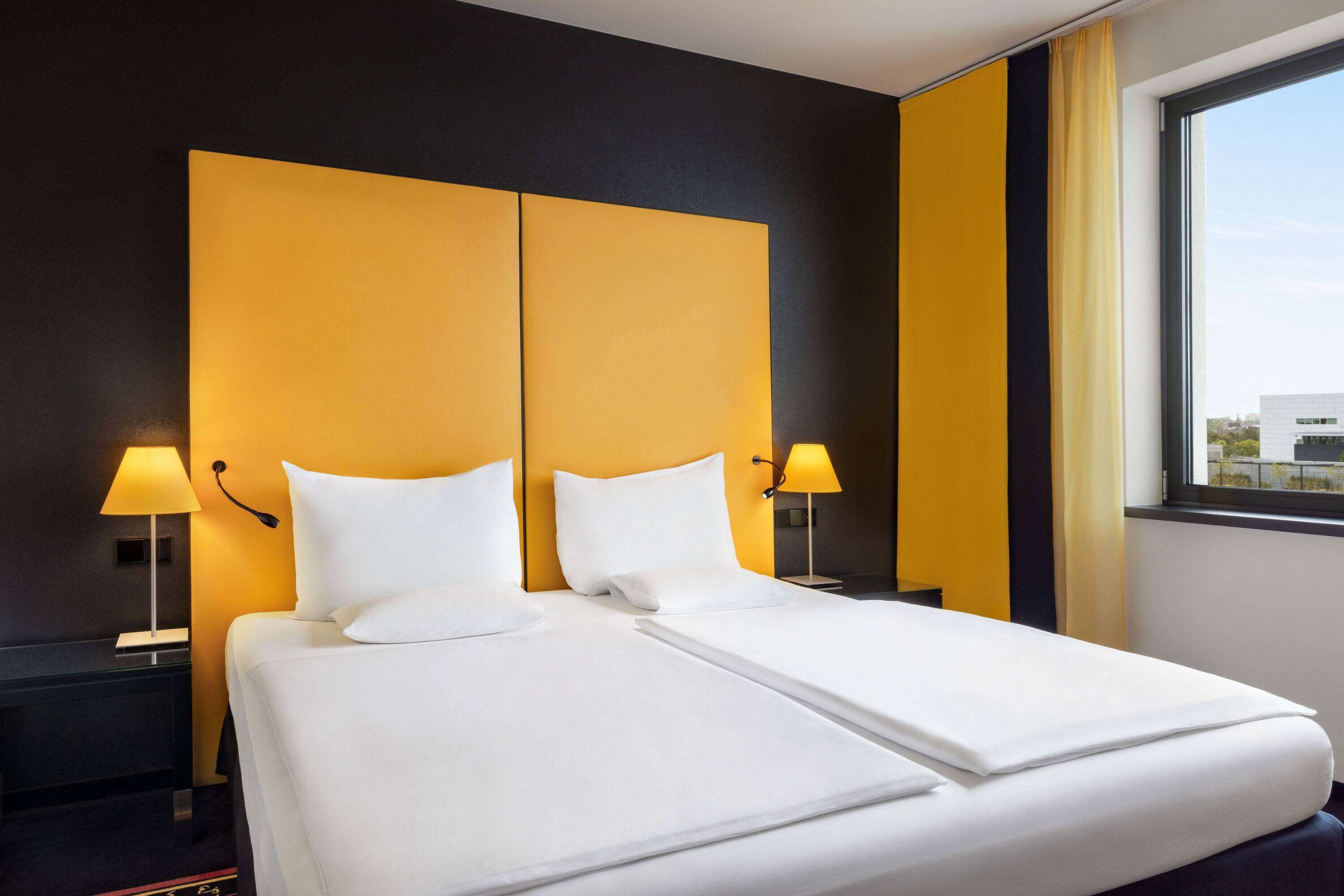 A hotel room with a large bed, two bedside lamps, and a view of the city through a window.